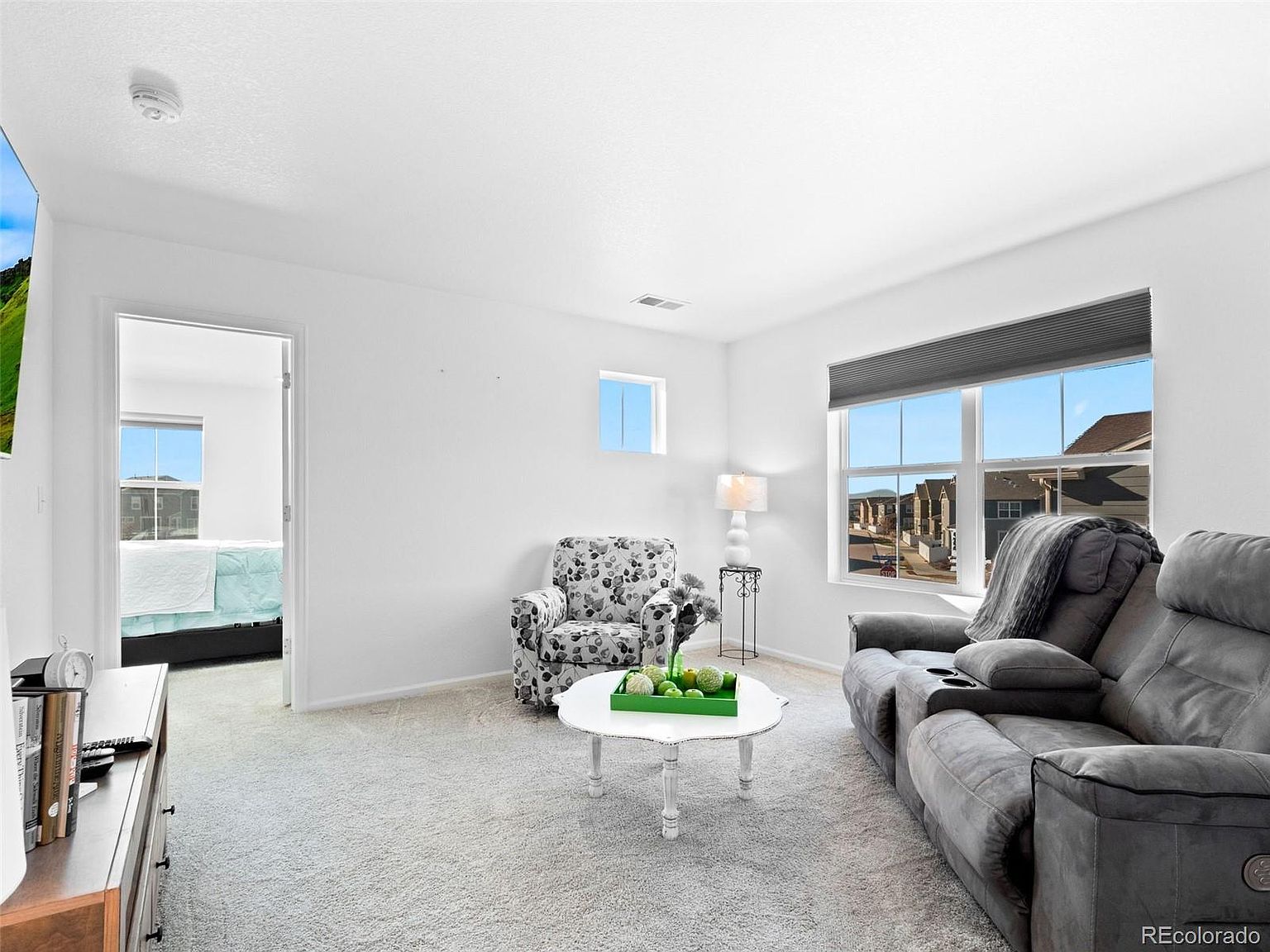 This is an interior shot of a living room featuring a gray reclining sofa, a floral armchair, and a white coffee table with decorative green spheres. Natural light floods the room through a large window, complemented by a smaller window on the adjacent wall. The room is carpeted and has white walls, creating a bright and airy atmosphere.