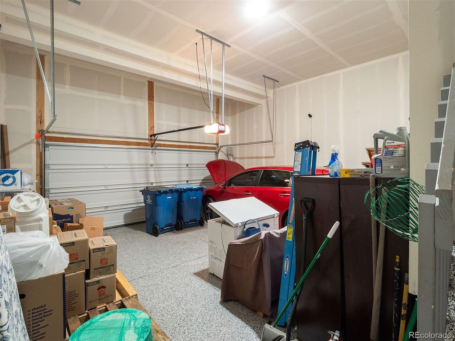 This is an interior shot of a garage. The garage features a white garage door, a red car parked inside, and several storage containers and boxes. The floor is a speckled gray, and the walls are painted in a neutral tone. The space appears to be well-lit and organized, though somewhat cluttered with various items.