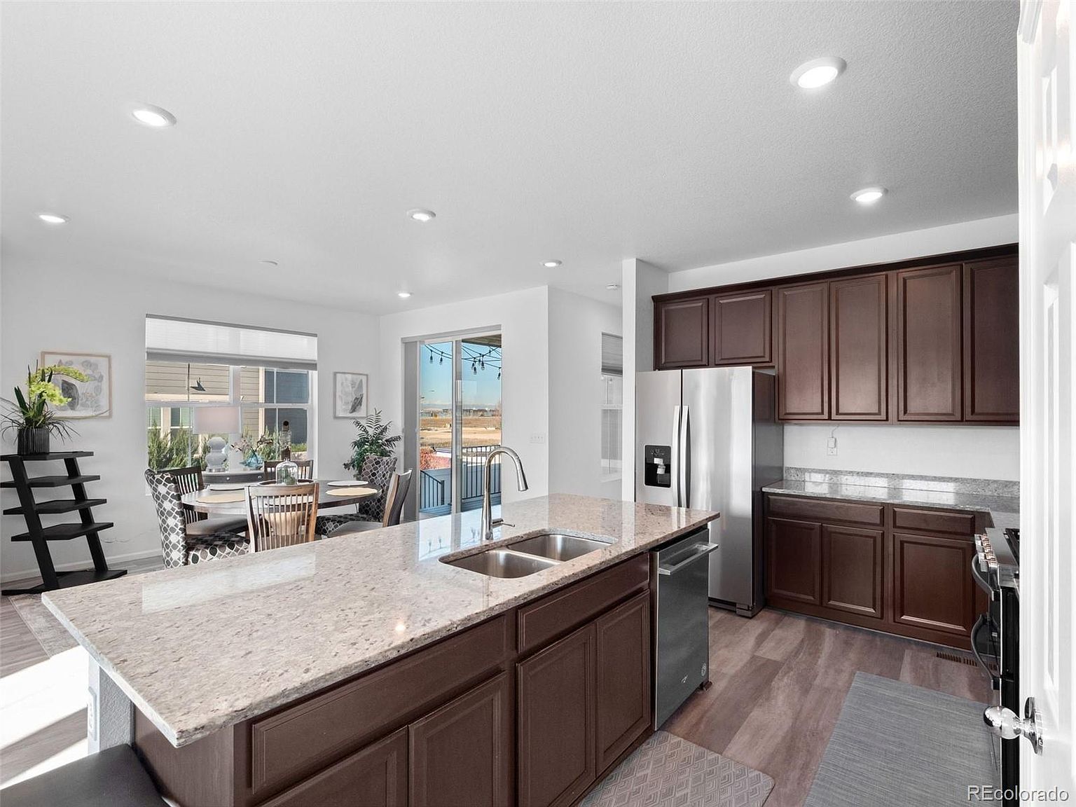 This is a well-lit kitchen featuring dark wood cabinetry and a large island with a light-colored granite countertop. Stainless steel appliances, including a refrigerator and dishwasher, are visible. The kitchen opens to a dining area with a view to the outside, creating an open and inviting space.