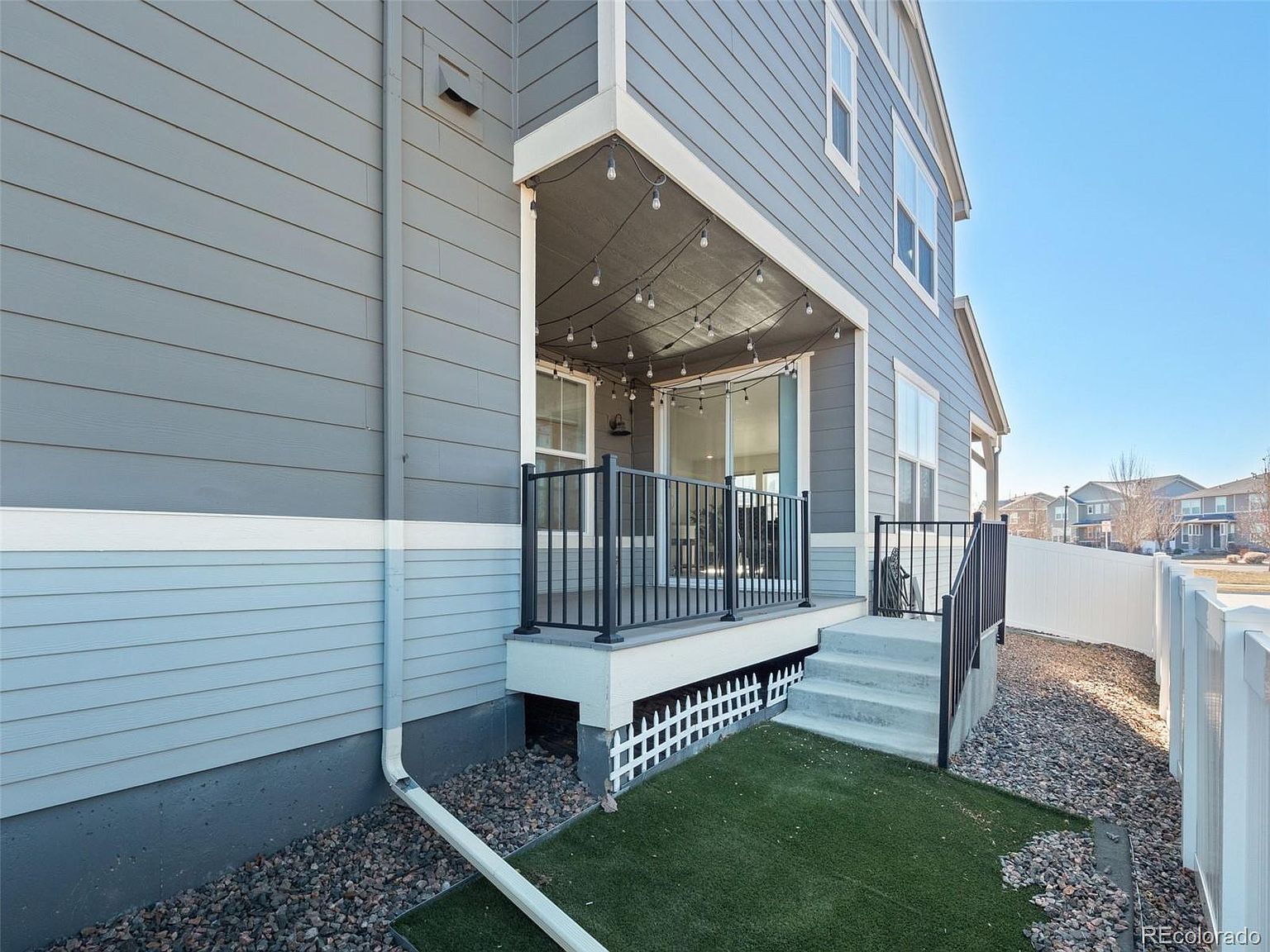 This image showcases a covered patio or deck area attached to a two-story home. The patio features a black metal railing, string lights hanging from the ceiling, and a sliding glass door leading inside. The exterior of the house is clad in gray siding with white trim, and there's a small patch of artificial turf adjacent to the patio steps, creating an inviting outdoor space.