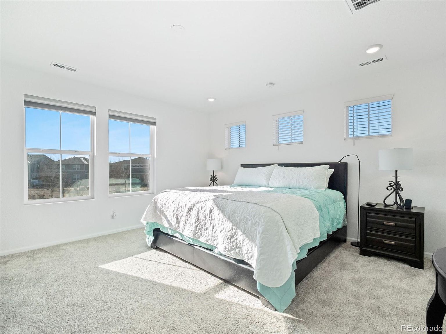This is a bright and airy primary bedroom featuring a large bed with a white comforter and light blue accents. The room has multiple windows that let in plenty of natural light, and the walls are painted a clean white. A dark wood nightstand with a lamp sits beside the bed, adding a touch of elegance.