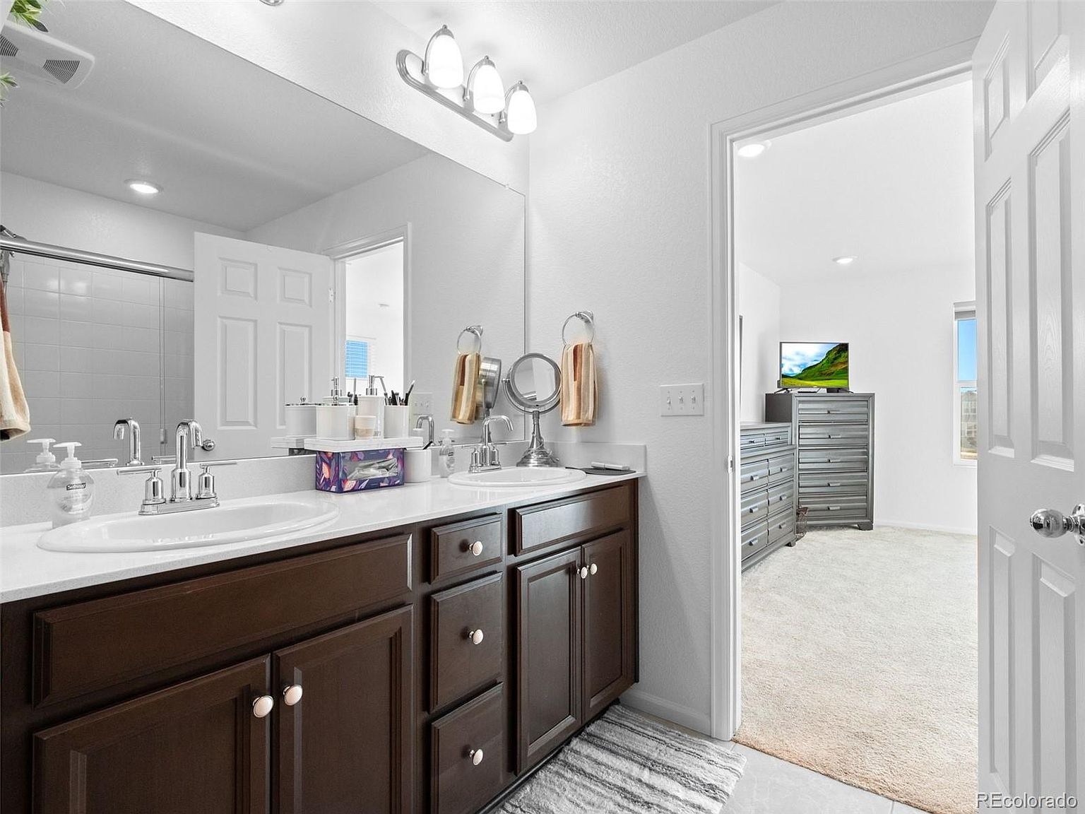 This is a well-lit primary bathroom featuring a double vanity with dark wood cabinets and white countertops. A large mirror spans the length of the vanity, reflecting the shower area. The bathroom opens into a bedroom with gray dressers and a television, creating a sense of spaciousness and connection.