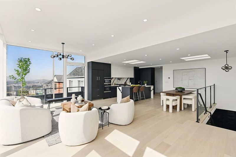 This is a bright and modern open-concept living space that seamlessly integrates the living room, kitchen, and dining area. The living room features comfortable white armchairs, a stylish coffee table, and large windows offering scenic views. The kitchen boasts sleek black and white cabinetry with a central island, while the dining area includes a contemporary table and chairs, creating an inviting and sophisticated atmosphere.
