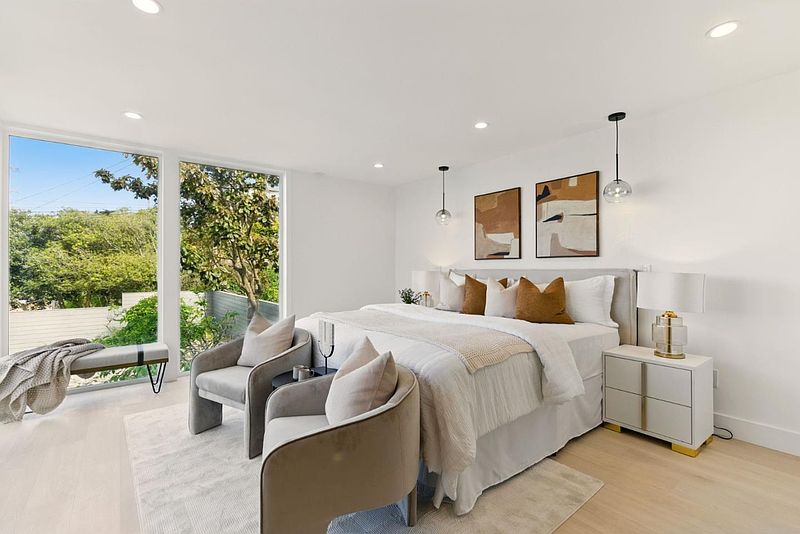 This is a bright and airy primary bedroom featuring a large window with a view of lush greenery. The room is decorated in neutral tones with modern artwork and stylish furniture, including two armchairs and a bench. The overall impression is one of comfort and sophistication.