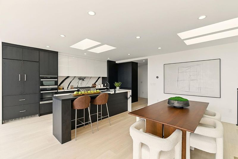 This is a modern kitchen with dark wood cabinetry and a contrasting white backsplash. The kitchen features an island with bar seating and stainless steel appliances. The dining area is adjacent to the kitchen, with a wooden table and unique white chairs, creating an open and inviting space.