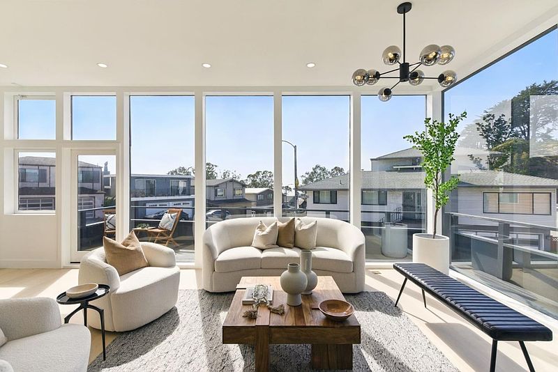 This is a bright and airy living room featuring floor-to-ceiling windows that offer an expansive view. The room is furnished with a modern curved sofa, comfortable armchairs, and a stylish coffee table, creating an inviting and sophisticated atmosphere. A contemporary chandelier adds a touch of elegance.