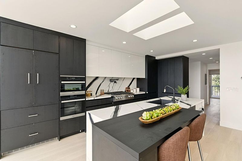 This modern kitchen features sleek, dark cabinetry contrasted with white upper cabinets and a striking marble backsplash. A large island with a dark countertop provides ample workspace and seating. Stainless steel appliances and recessed lighting contribute to the contemporary aesthetic, creating a sophisticated and functional space.