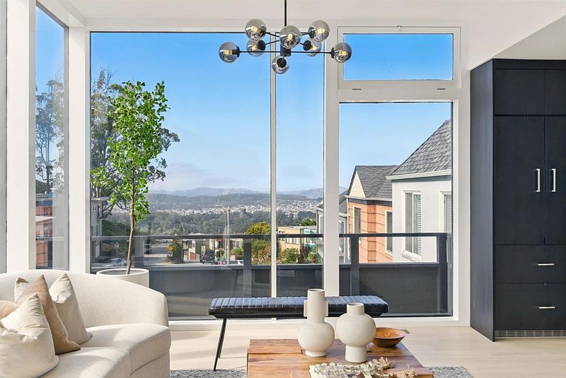 This is an interior shot of a living room featuring a large window with a view of the city. The room is furnished with a light-colored sofa, a bench, and a coffee table with decorative vases. A modern chandelier hangs from the ceiling, adding a touch of elegance to the space.