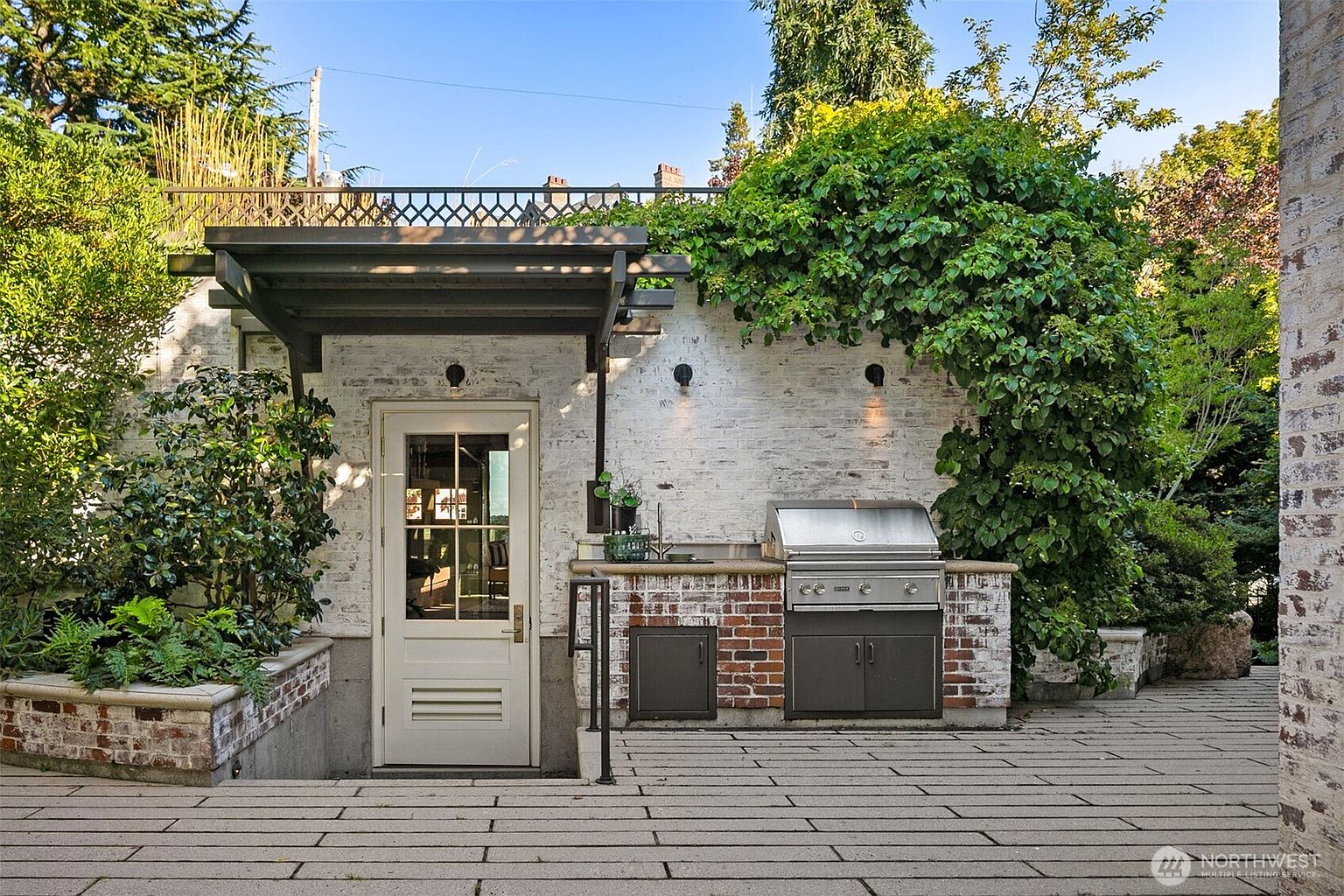 This image showcases an outdoor patio area featuring a built-in brick grill station with stainless steel appliances and storage. A white brick wall serves as a backdrop, partially covered by lush greenery, and a door provides access to the interior. The patio is paved with large rectangular stones, creating a clean and inviting outdoor living space.