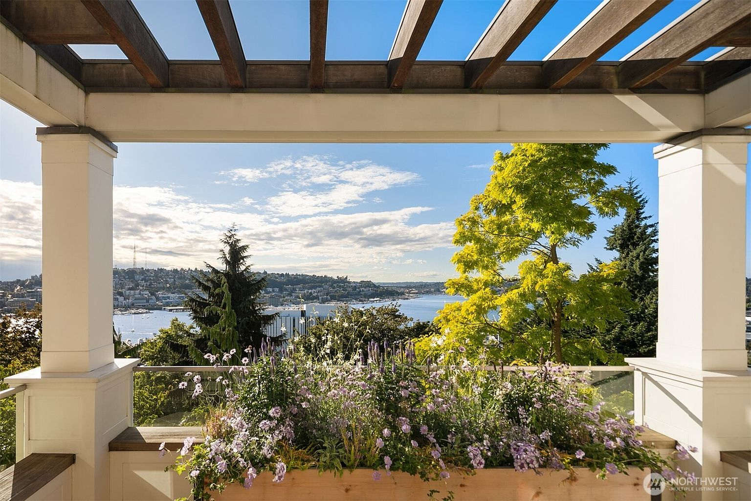 This image showcases a beautiful outdoor patio or balcony area with a stunning view. The space is framed by a wooden pergola and white pillars, with a planter box filled with purple flowers in the foreground. The background features a scenic view of a bay, city skyline, and lush greenery, creating a serene and inviting atmosphere.