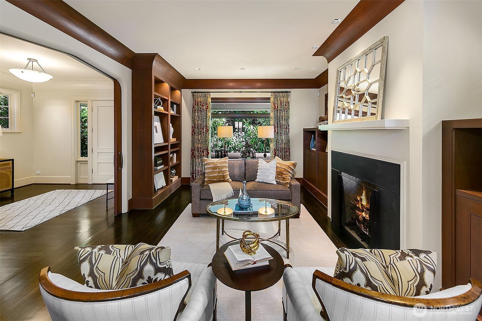 This is an interior shot of a living room featuring dark wood trim and built-in bookshelves. A neutral-toned sofa is centered in front of a window with patterned curtains, flanked by lamps. A fireplace with a decorative mirror above it adds a focal point, and two armchairs face the camera, creating an inviting and cozy atmosphere.