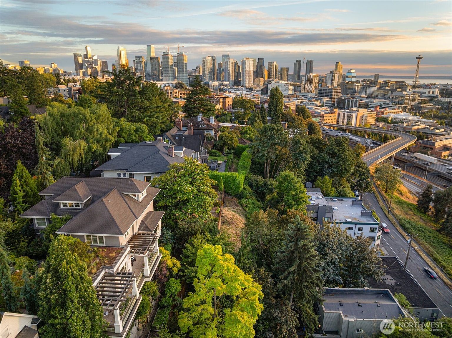 This aerial shot showcases a luxurious residential area with lush greenery and a stunning cityscape backdrop. Several multi-story homes with well-maintained roofs and balconies are nestled among mature trees. A highway with visible traffic adds a dynamic element to the scene, while the distant skyline provides a sense of urban proximity and prestige.