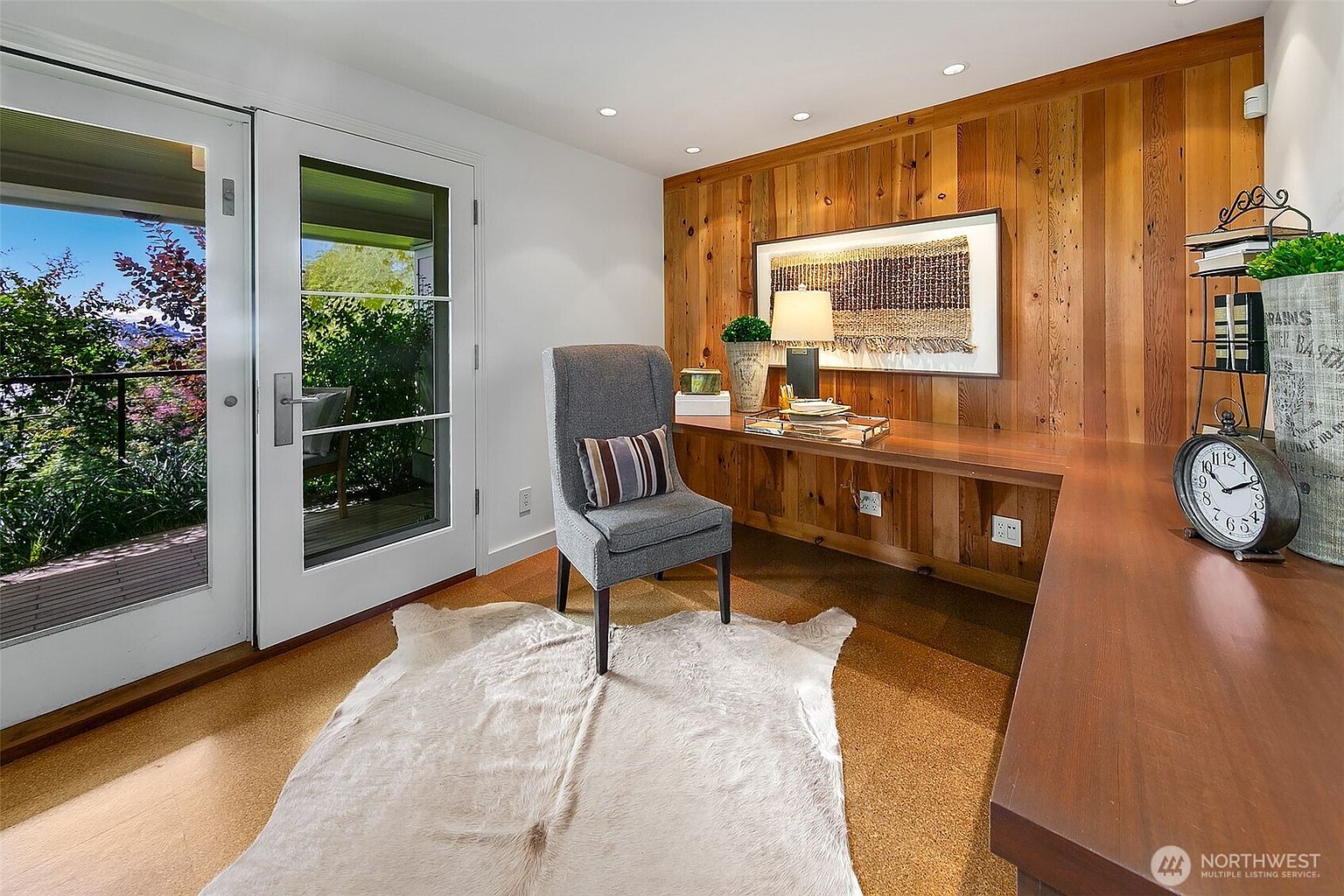 This is an interior shot of a home office or study. The room features a built-in wooden desk and wall paneling, complemented by a gray chair with a striped pillow. A white faux fur rug adds texture to the cork flooring, and double doors lead to an outdoor balcony with a view of lush greenery.