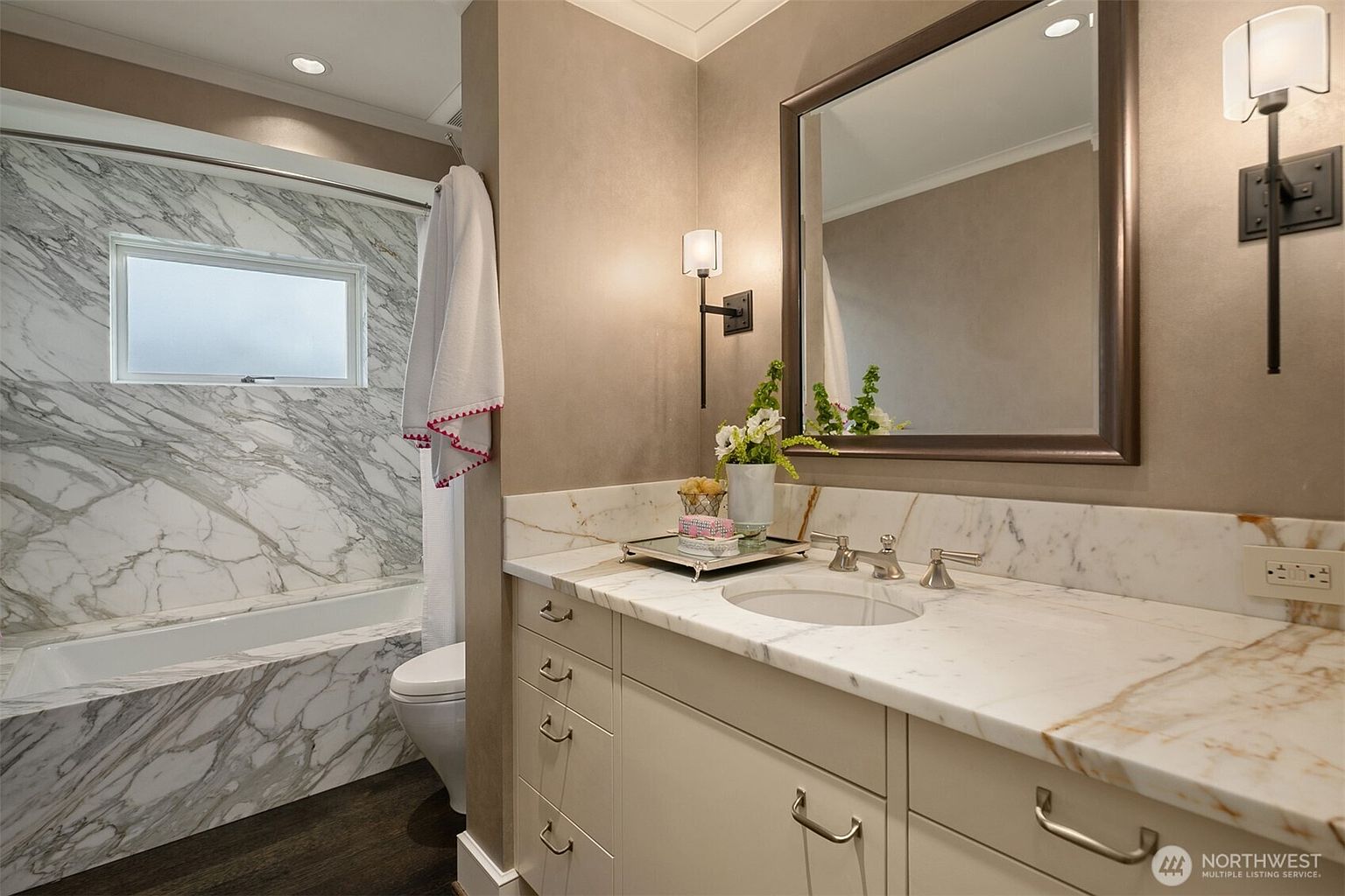 This is a well-lit bathroom featuring a marble-clad bathtub and shower combination, a toilet, and a vanity with a marble countertop. The vanity has light-colored cabinets with brushed nickel hardware. A framed mirror hangs above the vanity, and sconces provide additional lighting, creating a clean and elegant space.