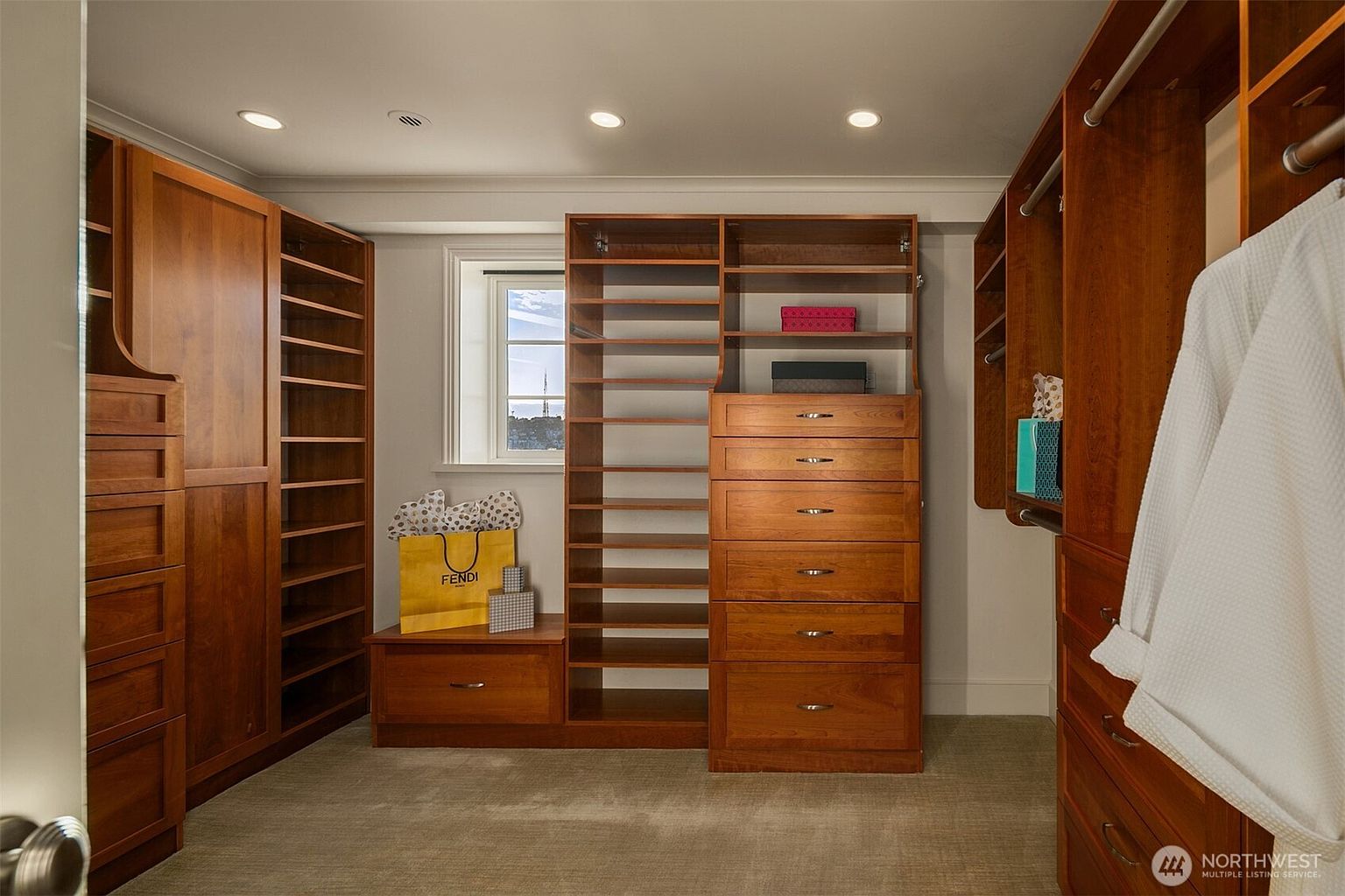 This is a well-organized walk-in closet featuring custom wood cabinetry and shelving. The closet includes drawers, open shelves, and hanging rods, providing ample storage space. A window allows natural light into the space, and a Fendi shopping bag adds a touch of luxury.