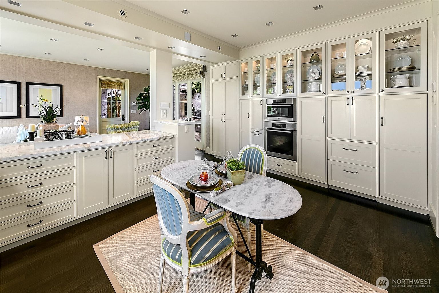 This is a well-lit kitchen featuring custom white cabinetry with glass-fronted display cabinets and built-in appliances. A marble-topped oval table with two chairs sits on a neutral rug, creating a cozy breakfast nook. The dark hardwood floors contrast with the light cabinetry, adding depth to the space.