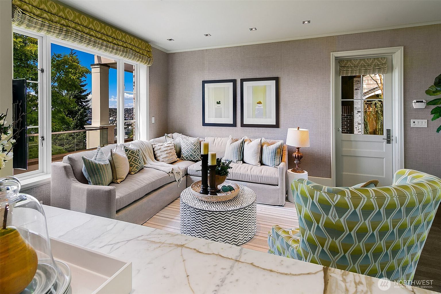 This is an interior shot of a living room featuring a large sectional sofa with decorative pillows, a patterned ottoman serving as a coffee table, and an accent chair with a geometric pattern. The room is well-lit with natural light from a large window offering a view of the outdoors, and the walls are adorned with framed artwork. The overall style is sophisticated and comfortable.