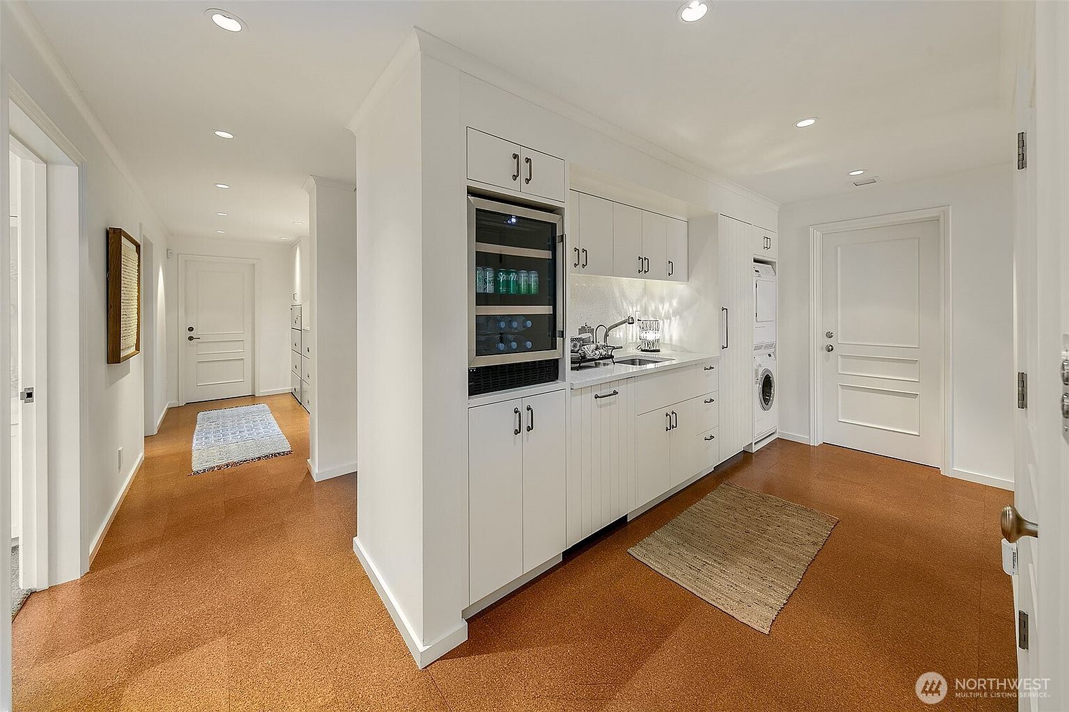 This interior shot showcases a well-organized laundry room with white cabinetry, a built-in wine cooler, and a stacked washer and dryer. The room features a sink with modern fixtures and a neutral-toned floor, creating a clean and functional space. A hallway extends from the laundry room, adding depth to the image.