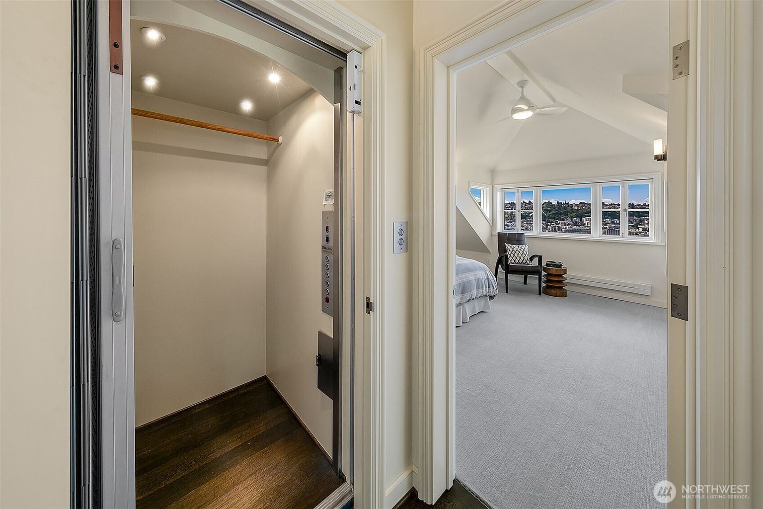 The image showcases a bright and airy bedroom with a view of the city skyline through a series of windows. The room features light gray carpeting, a bed with a striped comforter, and a modern chair. An elevator is visible in the foreground, adding a unique feature to the property.