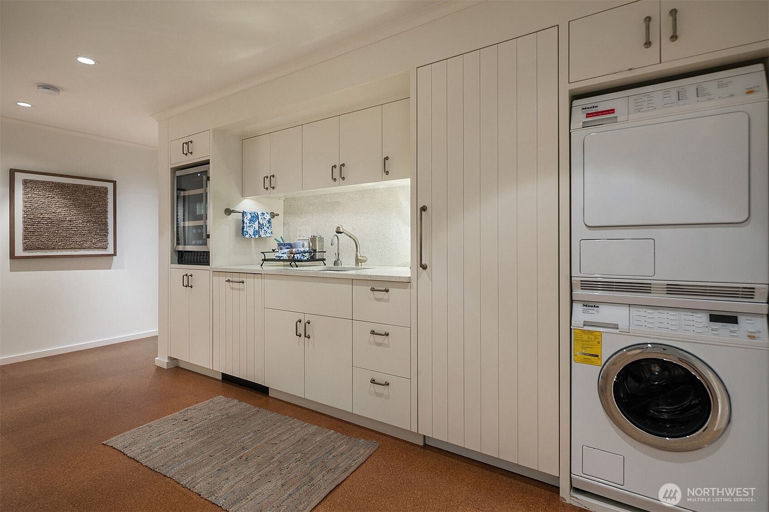 This is an interior shot of a laundry room featuring white cabinetry with bronze hardware, a sink, and stacked Miele washer and dryer. The room has a clean and organized appearance, with a neutral color palette and a modern aesthetic. A framed piece of art hangs on the wall, adding a touch of style to the space.