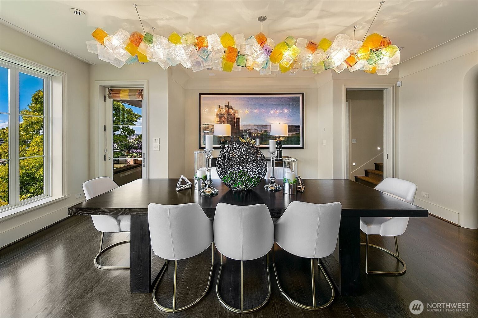 This is an interior shot of a dining room featuring a long, dark wood table surrounded by white chairs with gold accents. Above the table hangs a unique, colorful chandelier made of glass cubes, and a large framed artwork adorns the wall. The room is well-lit, creating an inviting and elegant atmosphere.
