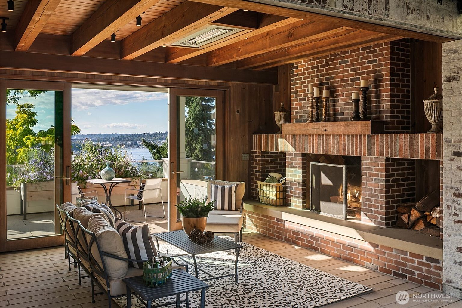 This sunroom features a brick fireplace, wood-paneled walls and ceiling, and sliding glass doors that open to a patio with a view of the water. The room is furnished with a sofa, chairs, and tables, creating a cozy and inviting space. The overall impression is one of warmth and relaxation, perfect for enjoying the natural light and outdoor scenery.