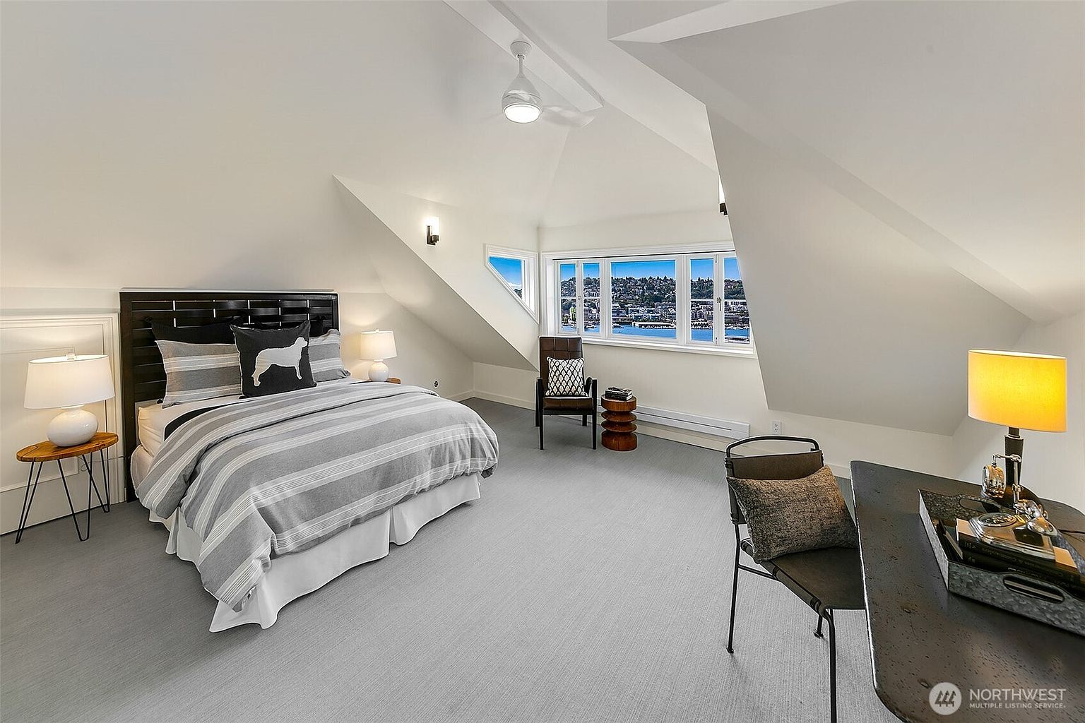 This is a bright and airy primary bedroom featuring a bed with a striped duvet and decorative pillows, a dark wood headboard, and a side table with a lamp. The room has a sloped ceiling, a ceiling fan, and a window with a view of the water. A desk and chair are also present, creating a functional and stylish space.