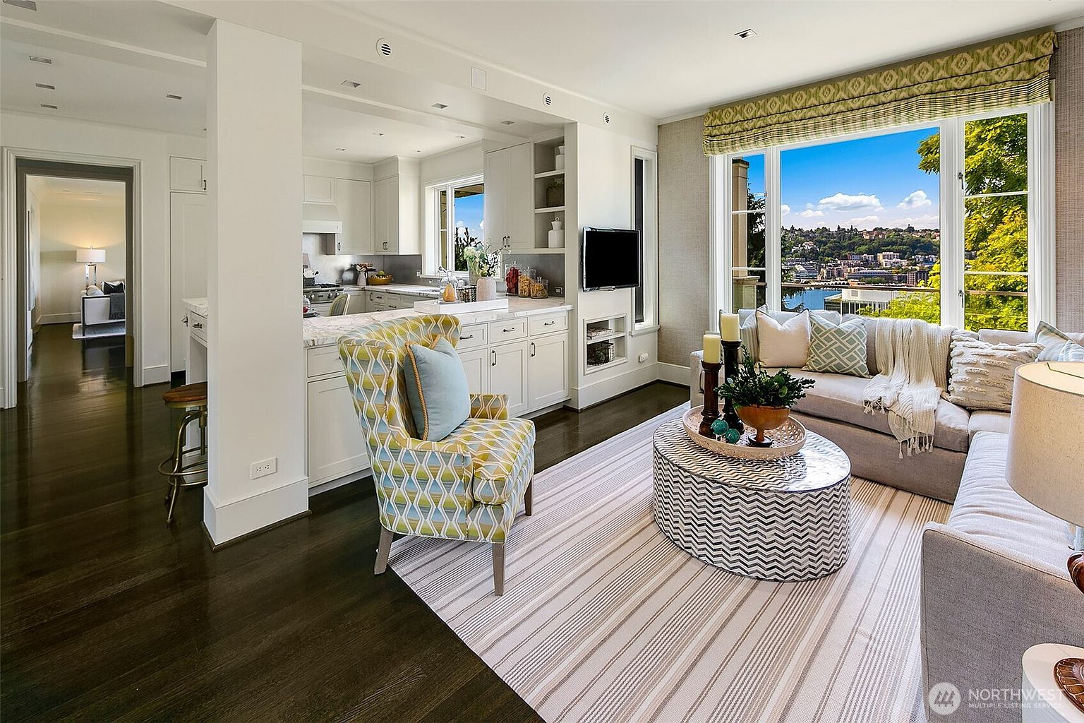 This is an interior shot of a living room that seamlessly blends into a kitchen area. The living room features a comfortable seating arrangement with a sofa, patterned armchair, and a unique coffee table, all set on a striped rug. Large windows offer a scenic view, enhancing the room's appeal, while the kitchen area is visible in the background, showcasing white cabinetry and countertops.
