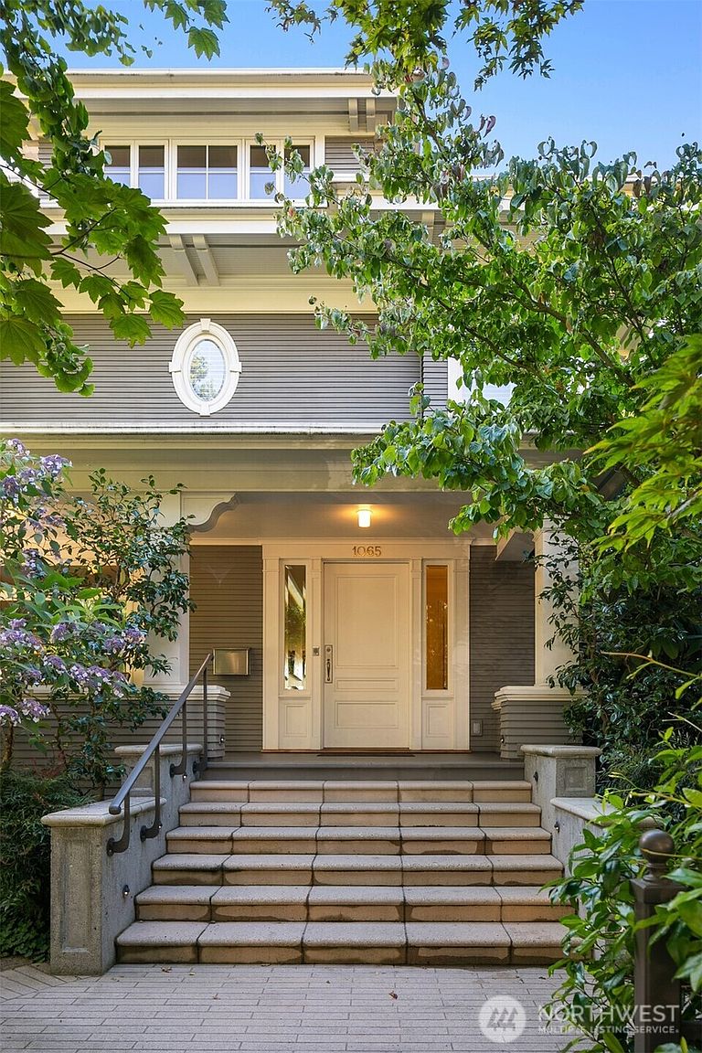 This image showcases the inviting entryway of a home, featuring a set of stone steps leading up to a white front door with sidelights. The house has a gray exterior with an oval window above the porch. Lush greenery frames the entrance, adding to the curb appeal and creating a welcoming atmosphere.