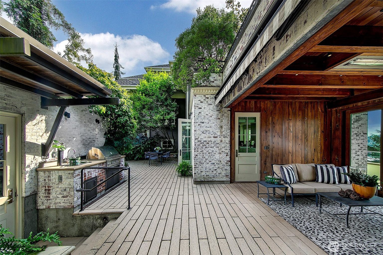 This image showcases a beautifully designed outdoor patio area. The space features a built-in outdoor kitchen with a grill, a comfortable seating area with a sofa and coffee table, and a spacious deck made of light-colored pavers. The patio is partially covered, providing shade and protection from the elements, and is surrounded by lush greenery, creating a private and inviting atmosphere.