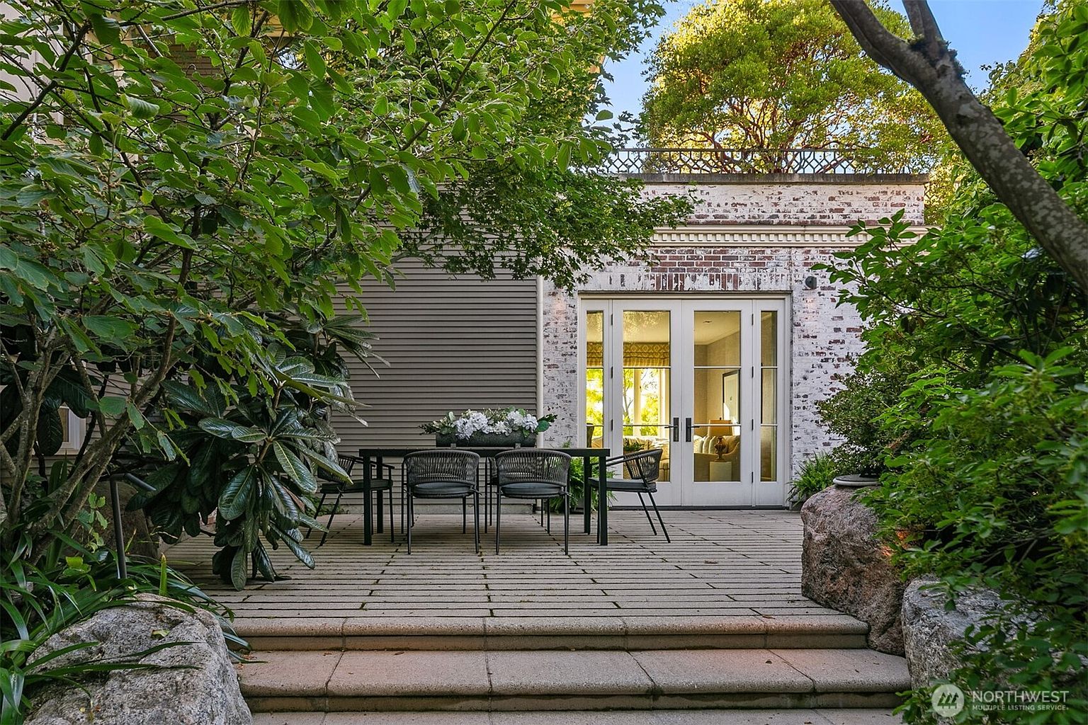 This image showcases a charming outdoor patio area, featuring a dining set with a black table and woven chairs, set upon a stone-tiled surface. The patio is surrounded by lush greenery and mature trees, creating a private and inviting atmosphere. The backdrop includes a building with a brick facade and large glass doors, suggesting a seamless transition between indoor and outdoor living spaces.