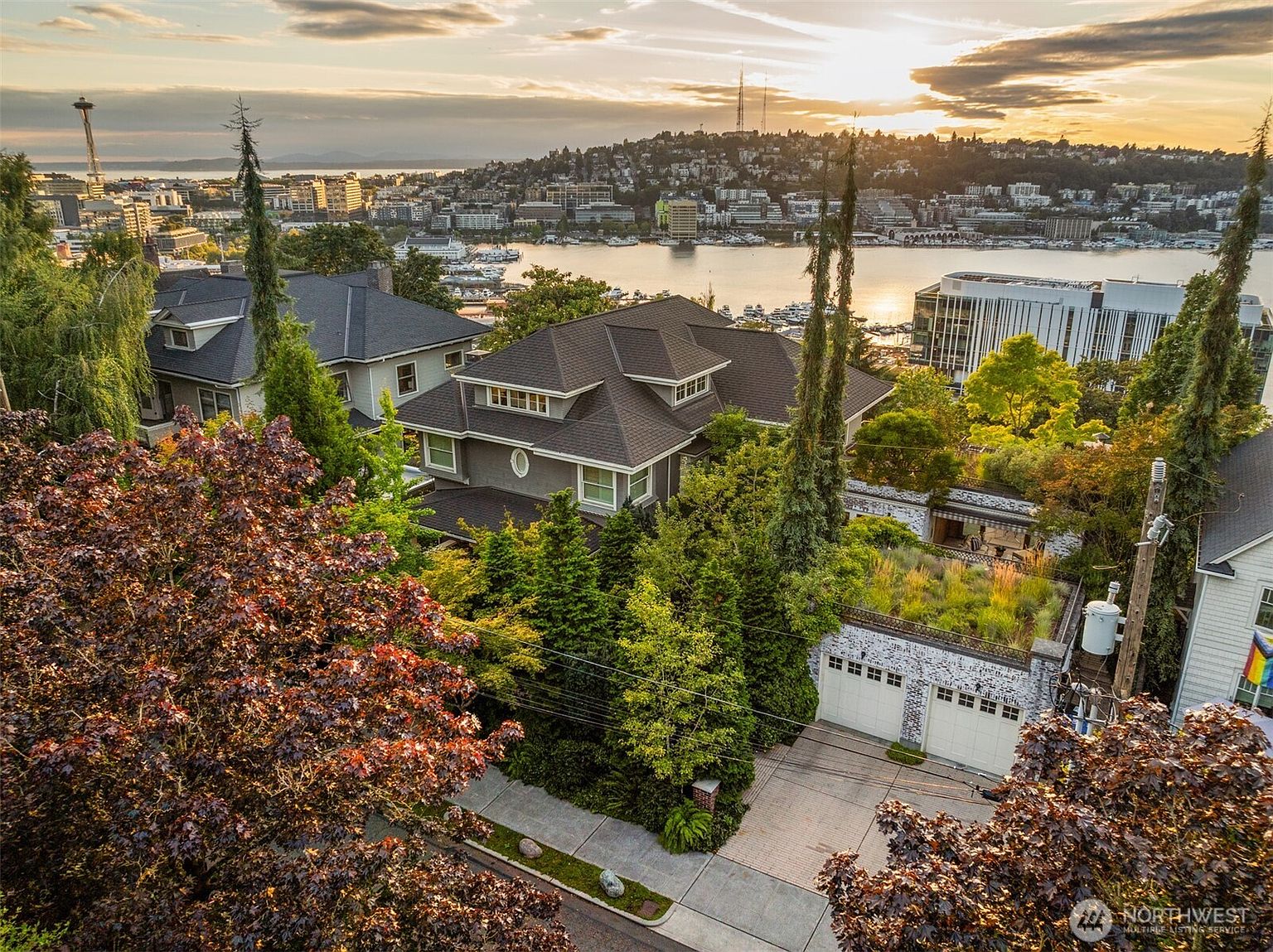 This aerial shot showcases a beautiful multi-story home with a dark roof, surrounded by lush greenery and mature trees. The property features a unique garage with a green roof, adding to its appeal. In the background, a stunning cityscape and waterfront view enhance the property's desirability, creating a sense of luxury and exclusivity.