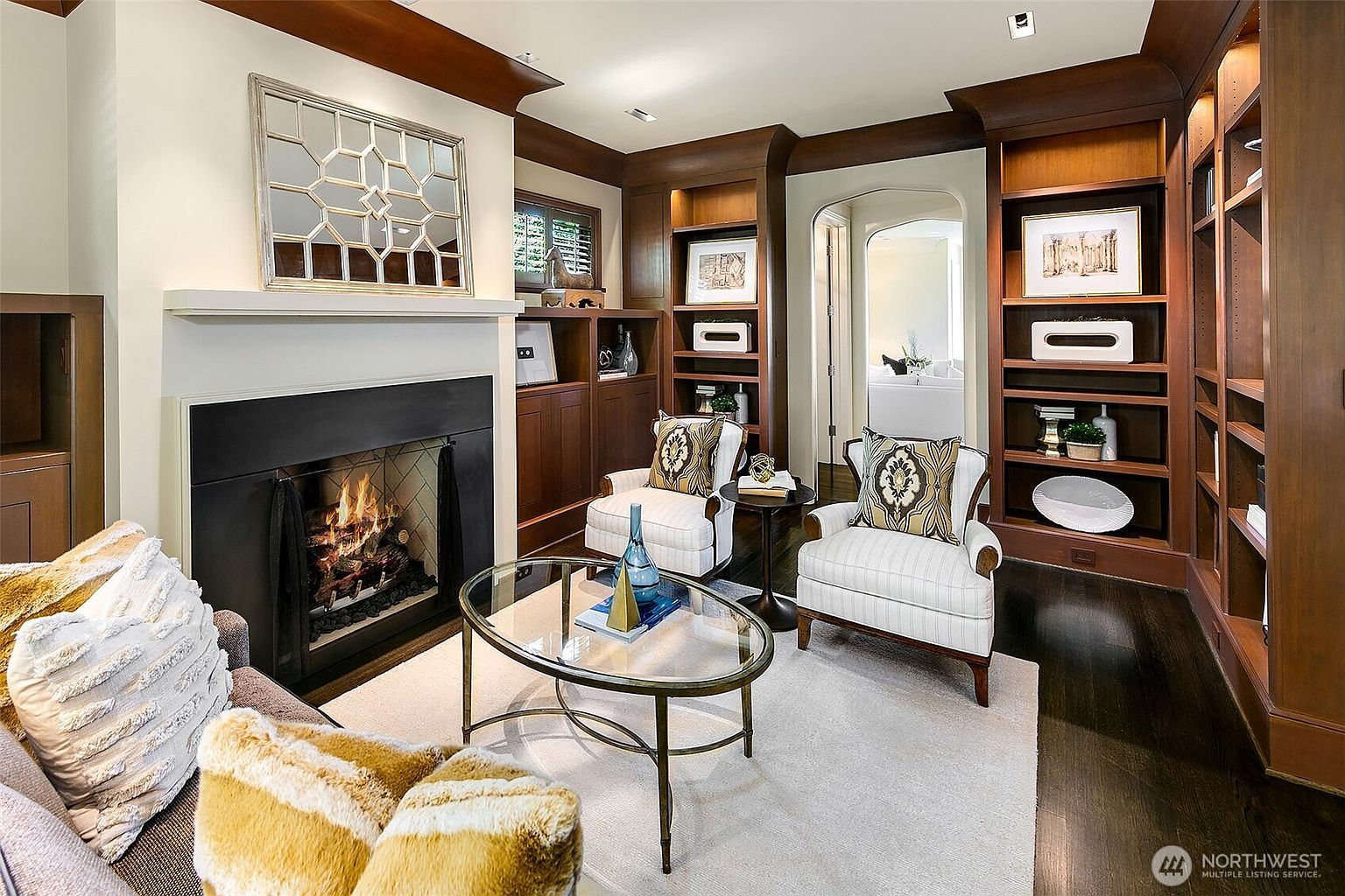 This is an inviting living room featuring a fireplace with a decorative mirror above, flanked by built-in wooden shelving units. Two armchairs face a glass coffee table, set on a light-colored rug. The room exudes a cozy and sophisticated ambiance, perfect for relaxation and entertaining.