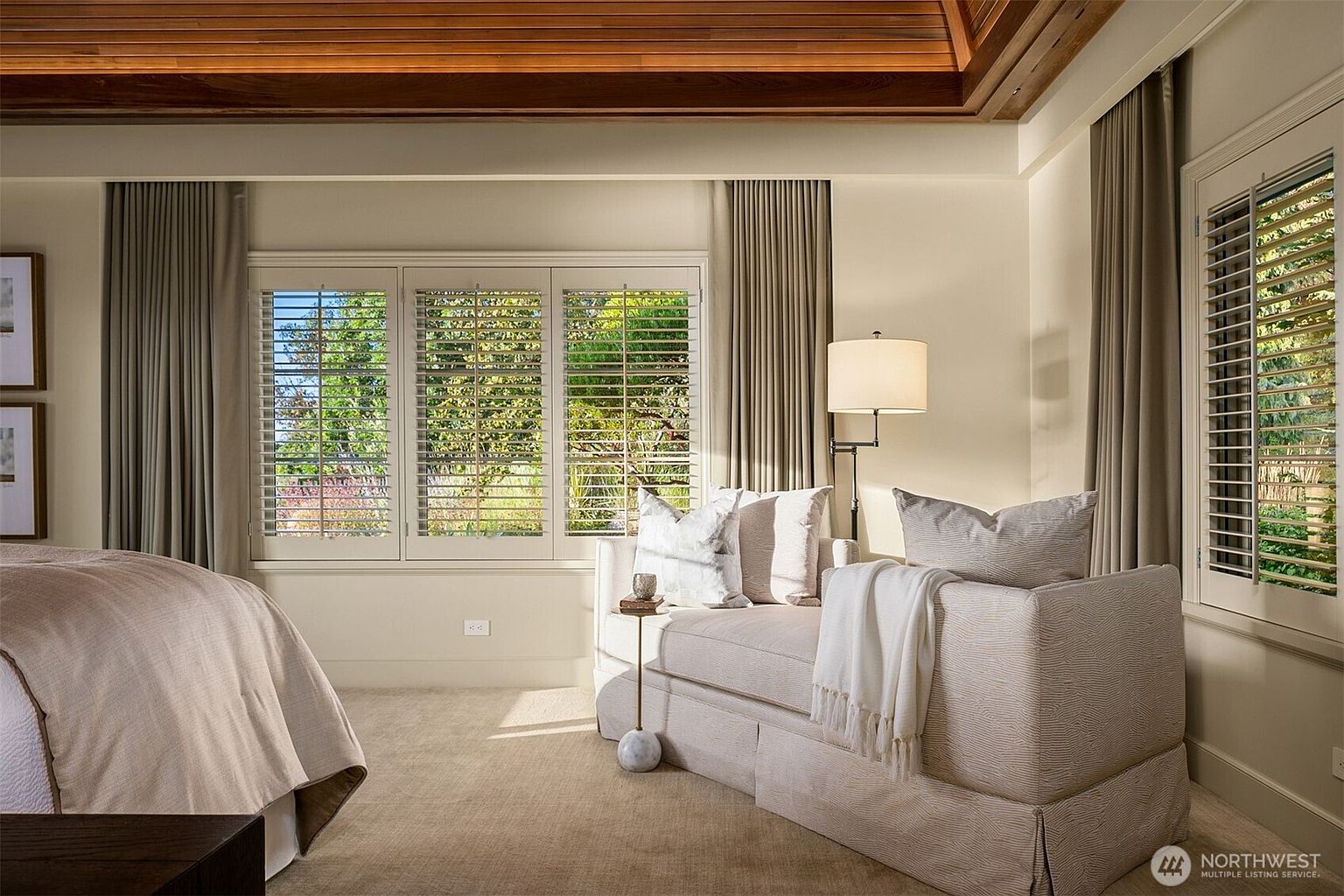 This is a well-lit primary bedroom featuring a seating area with a comfortable sofa, complemented by neutral-toned curtains and plantation shutters on the windows. The room has a serene and inviting atmosphere, with a glimpse of a bed on the left side, suggesting a spacious layout. The wooden ceiling adds a touch of warmth and character to the space.