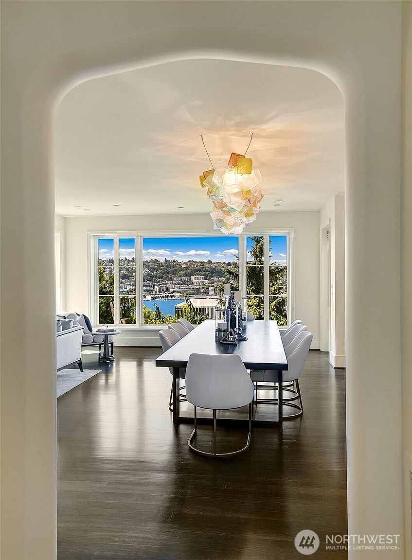 This is an interior shot of a dining room featuring a long, dark wood table with light-colored chairs. A modern, colorful chandelier hangs above the table, and a large window provides a scenic view of a cityscape and water. The room is accessed through an arched doorway, adding an elegant touch to the space.