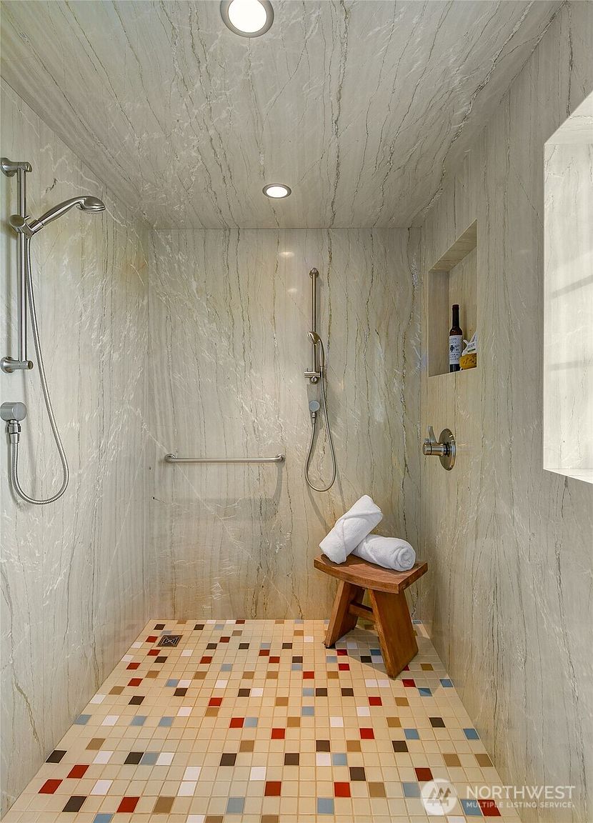 This is a well-lit primary bathroom featuring a walk-in shower with marble-like walls and a mosaic tile floor. The shower includes both a fixed showerhead and a handheld sprayer, along with a grab bar for accessibility. A small wooden stool with rolled towels and a niche with decorative items add a touch of elegance to the space.