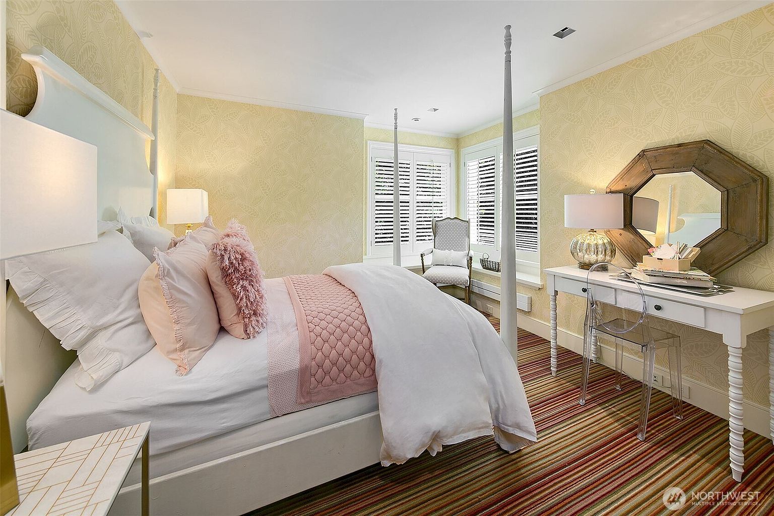 This is a bedroom featuring a four-poster bed with white linens and pink accents, including pillows and a quilted throw. The room has patterned yellow wallpaper and a white desk with a mirror above it, complemented by a clear acrylic chair. The flooring is a striped rug, adding a touch of color and texture to the space.