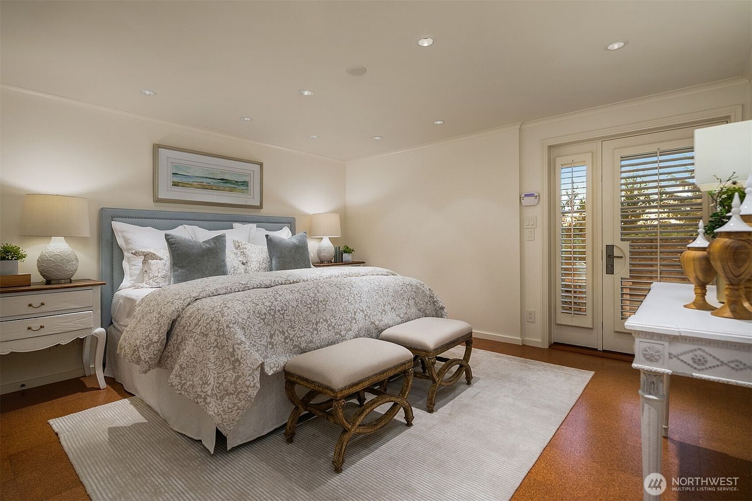 This is a well-lit primary bedroom featuring a large bed with a patterned duvet and several pillows. The room includes a bedside table with a lamp, a pair of upholstered benches at the foot of the bed, and a white table with decorative items near a set of French doors. The overall impression is one of comfort and understated elegance.