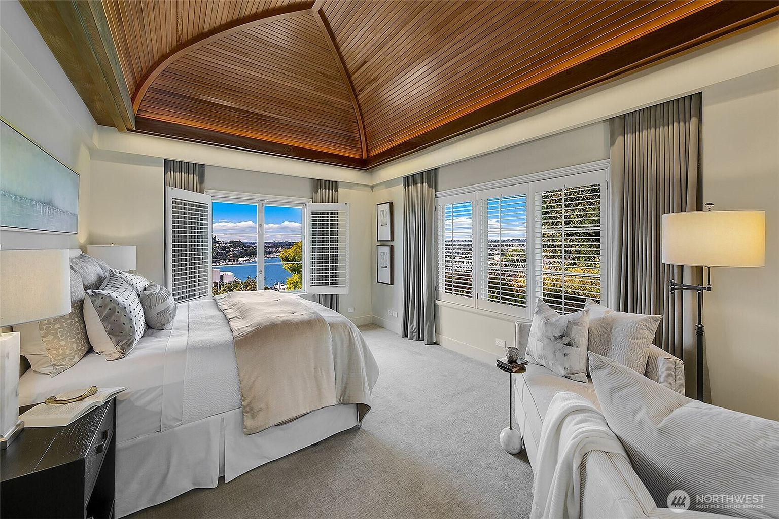 This is a primary bedroom featuring a large bed with white linens and decorative pillows, positioned to take advantage of the scenic views from the windows. The room includes a seating area with a sofa and a side table, complemented by elegant gray curtains and a unique wooden ceiling design. The overall impression is luxurious and serene, emphasizing comfort and style.