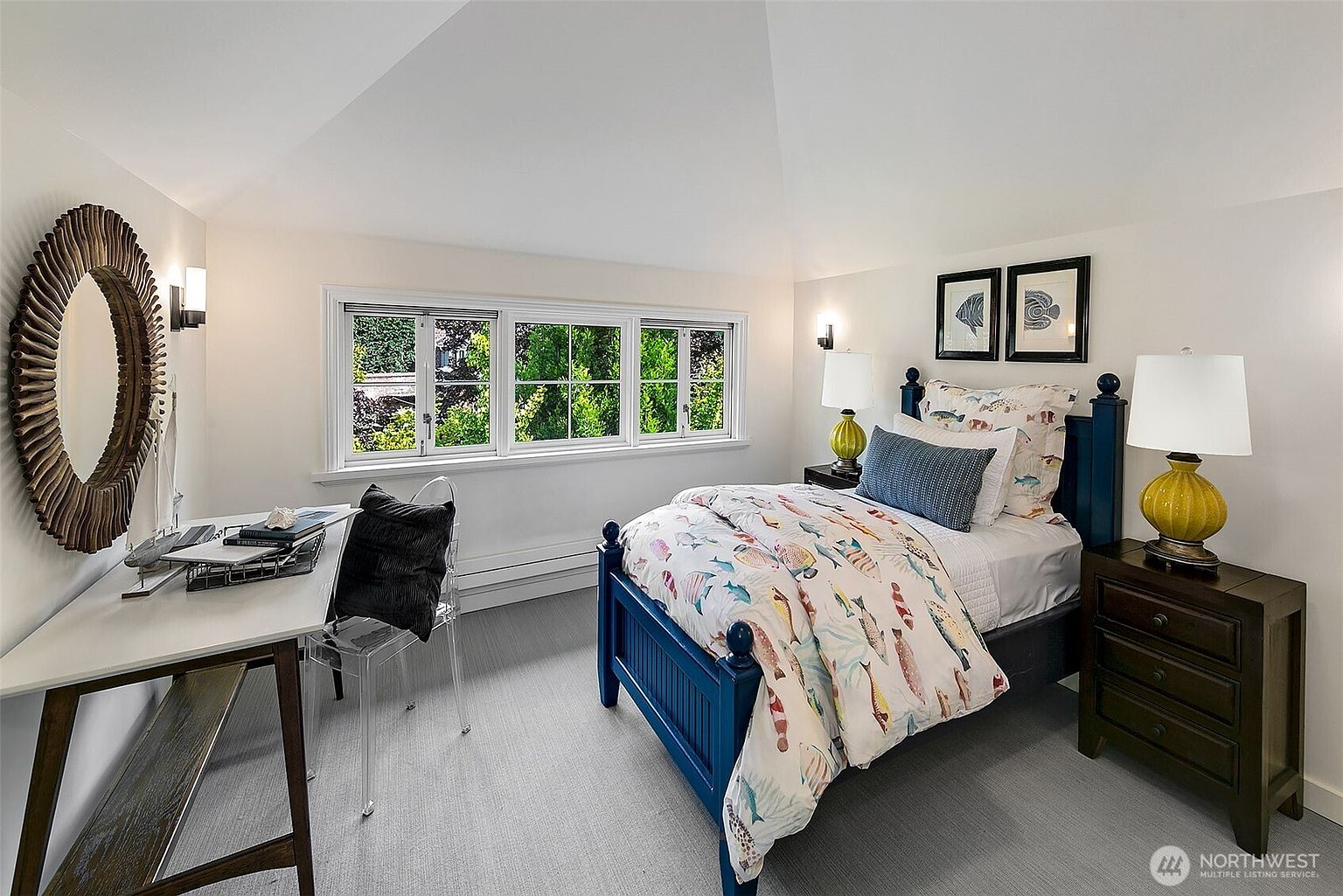 This is a bright and inviting guest bedroom featuring a blue-painted bed with a whimsical fish-themed duvet cover. The room is well-lit with natural light from a window overlooking greenery, complemented by two table lamps on either side of the bed. A desk area with a unique round mirror adds a functional and stylish touch to the space.