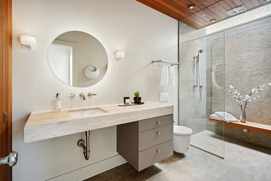 This modern bathroom features a floating vanity with a light-toned stone countertop and a circular backlit mirror. To the right, a glass-enclosed walk-in shower showcases textured stone wall tiles and a built-in wooden bench. The space is accented by a warm wood-paneled ceiling and neutral-toned floor tiles, creating a serene and spa-like atmosphere.