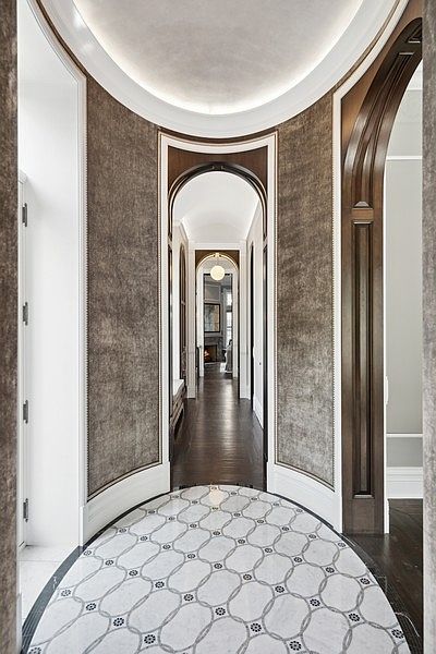This elegant hallway features a curved entryway with a sophisticated patterned stone floor and textured wall coverings. A series of arched doorways with dark wood trim create a sense of depth and architectural rhythm, leading the eye toward a distant room with a fireplace. The perspective is centered and symmetrical, emphasizing the grand, transitional nature of the home's layout.