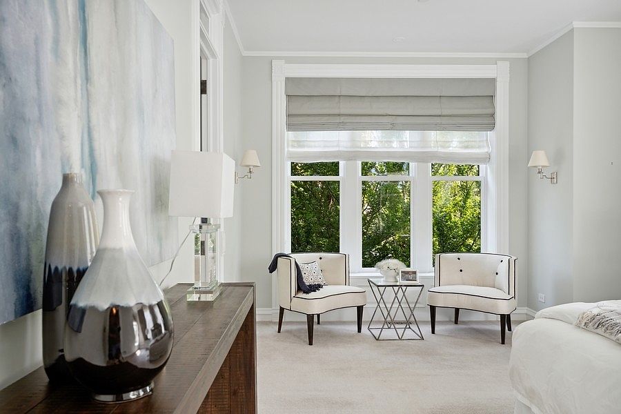 This elegant bedroom features a cozy seating area positioned in front of a large window with soft gray Roman shades. The space is furnished with two cream-colored armchairs, a small geometric metal side table, and a wooden console table adorned with decorative vases and a lamp. The neutral color palette, plush carpeting, and wall sconces create a serene and sophisticated atmosphere.
