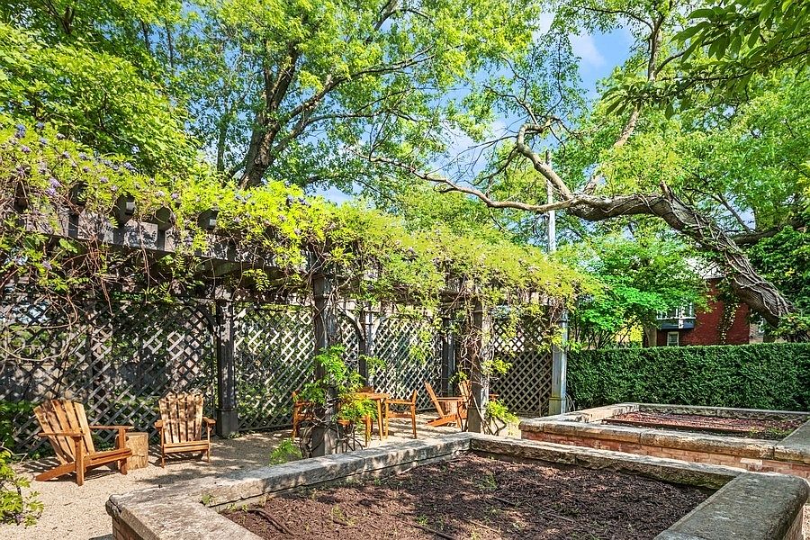 This serene garden space features a large wooden pergola covered in climbing vines, providing a shaded seating area with rustic wooden chairs. In the foreground, two raised stone garden beds filled with dark soil anchor the space, while a gravel path winds through the area. The scene is framed by lush green trees and a manicured hedge, creating a private and tranquil outdoor retreat.
