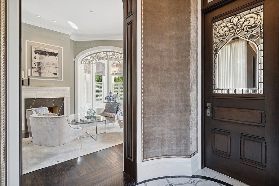 This view from a foyer looks through a wide, arched doorway into a sophisticated living room. The foreground features a dark wood door with an intricate leaded glass insert, while the living area beyond showcases a fireplace with a stone surround, plush seating, and large windows with elegant window treatments. The transition between spaces is defined by rich, dark wood flooring and a curved wall finished in a textured, neutral-toned material.