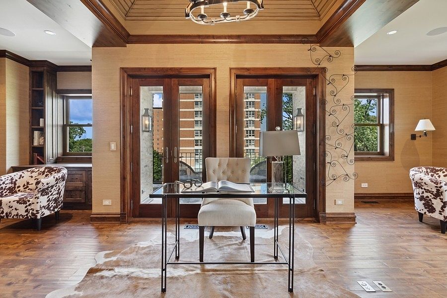This sophisticated home office features a central glass-topped desk paired with a tufted upholstered chair, set against a backdrop of double French doors that open to a balcony. The room is anchored by a large animal-hide rug on dark wood flooring, complemented by two cow-print armchairs and custom built-in shelving. The space is illuminated by a decorative chandelier and a table lamp, creating a warm and professional atmosphere.