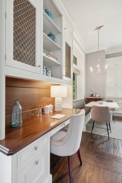 This elegant home office space features a built-in desk with a rich wood surface and white cabinetry, including open shelving and lattice-fronted upper cabinets. A tufted upholstered chair sits at the desk, which is illuminated by a textured table lamp, while a dining area with a chandelier and upholstered chairs is visible in the background. The room is finished with dark wood flooring laid in a herringbone pattern, creating a sophisticated and cohesive aesthetic.