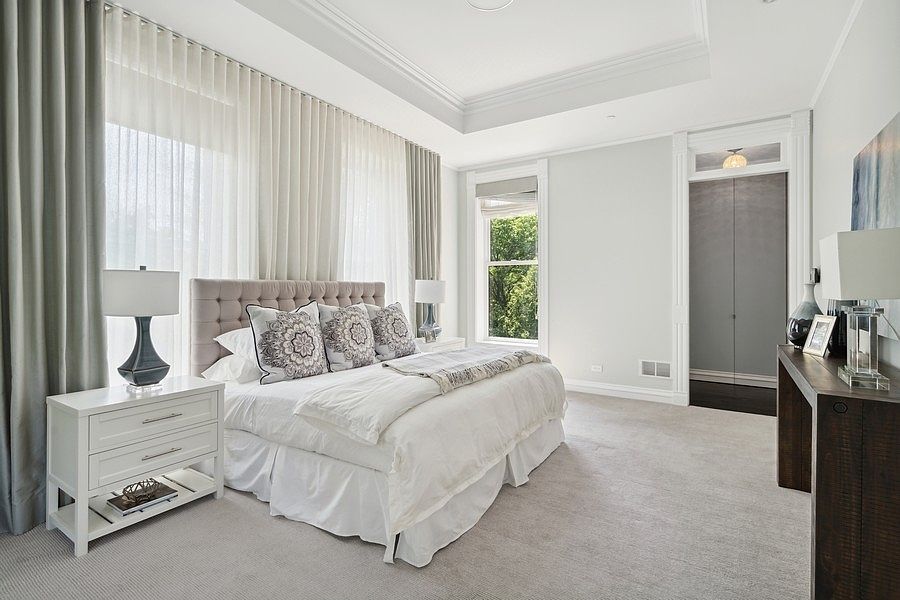 This elegant primary bedroom features a plush, tufted headboard centered against a wall of floor-to-ceiling sheer curtains that soften the natural light. The room is appointed with a white nightstand, a dark wood console table, and a neutral color palette that creates a serene and sophisticated atmosphere. A tray ceiling adds architectural depth, while the clean lines and soft textures provide a luxurious, restful aesthetic.