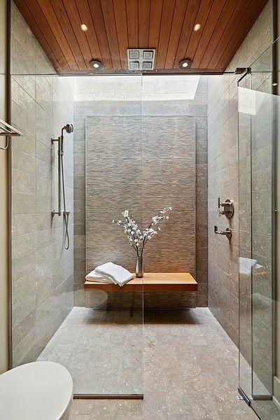 This luxurious walk-in shower features a striking textured stone accent wall and a floating light wood bench adorned with a decorative vase of flowers. The space is enclosed by clear glass panels and topped with a warm wood-slat ceiling, creating a spa-like atmosphere. The perspective is a direct, eye-level shot looking into the shower stall, highlighting the clean lines and high-end finishes.