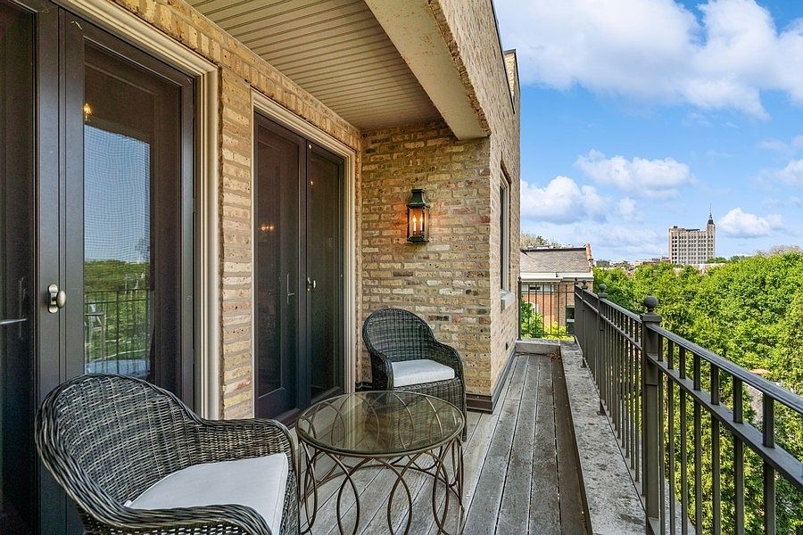 This elevated balcony features a weathered wood deck and a classic brick exterior wall, creating a cozy outdoor retreat. Two dark wicker chairs with white cushions are arranged around a small glass-topped table, perfect for enjoying the scenic view of the surrounding trees and distant city skyline. The space is illuminated by a traditional wall-mounted lantern, offering a charming and inviting atmosphere.